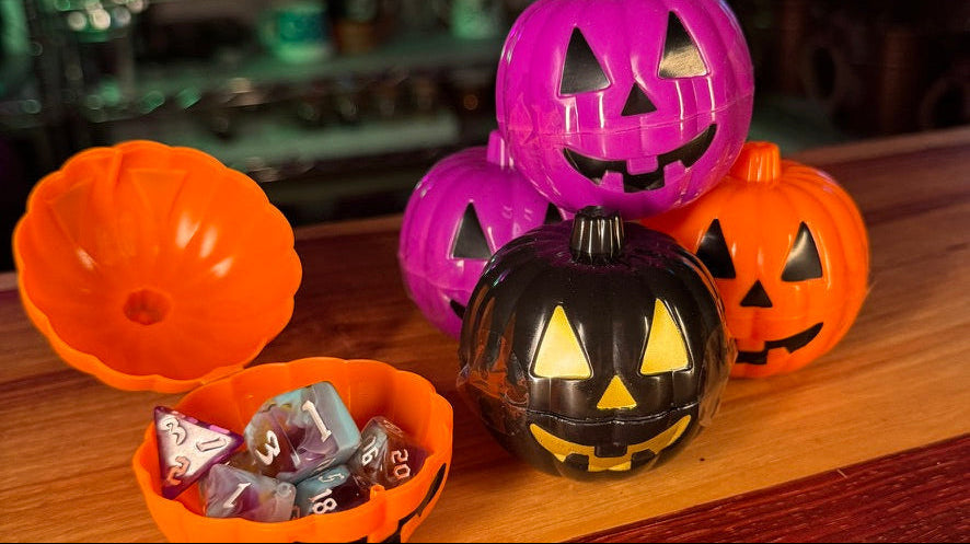 A set of Halloween-themed plastic storage containers shaped like jack-o'-lanterns, including an orange lid, an orange container filled with dice, and three pumpkins in black, purple, and orange holding candies, arranged on a wooden tabletop.